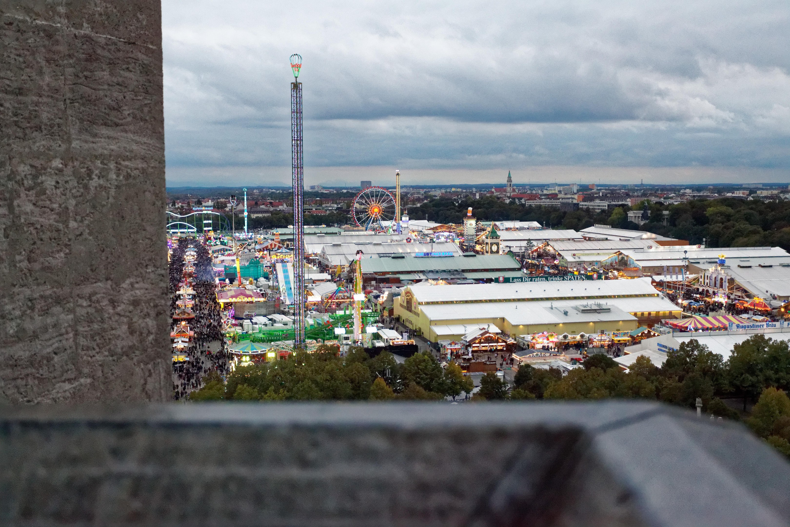 Sicht auf einen Vergnügungspark von oben, mit einer Wand, Bäumen, Gebäuden, Fahrgeschäften und Pfählen im Vordergrund und einem bewölkten Himmel im Hintergrund.