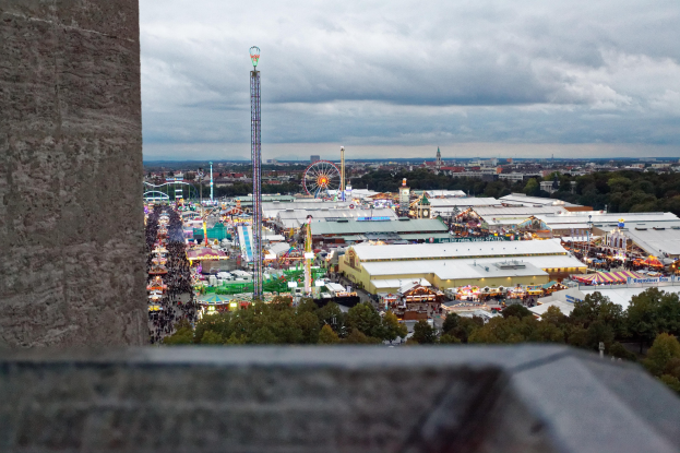 Sicht auf einen Vergnügungspark von oben, mit einer Wand, Bäumen, Gebäuden, Fahrgeschäften und Pfählen im Vordergrund und einem bewölkten Himmel im Hintergrund.