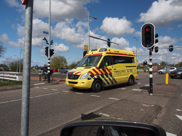 Ambulanz fährt auf einer Straße neben einer Ampel, mit einer Person auf einem Fahrrad im Vordergrund, umgeben von Laternenmästen, Bäumen, Gebäuden und einem bewölkten Himmel, und ein Spiegel ist unten sichtbar.