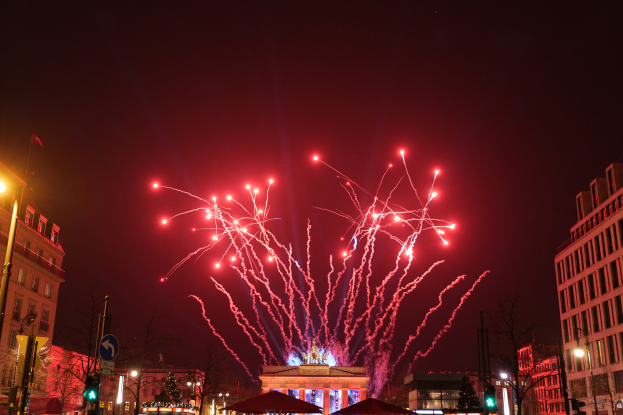Eine Stadtstraße in Berlin an Silvester, voller Gebäude, Bäume, Laternenpfähle, Verkehrszeichen, Zelte und Menschen, mit einem spektakulären Feuerwerk, das den Himmel erhellt.