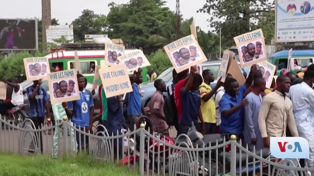 Eine Gruppe von Menschen mit Schildern, die vor einem Zaun protestieren, umgeben von Fahrzeugen, B├Ąumen, Masten, Plakaten und einem Bildschirm, mit Himmel und Gras im Hintergrund.