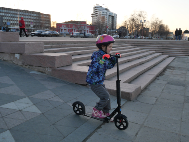 Ein junges Mädchen in einem Helm fährt auf einem Scooter eine Treppe hinunter mit Fahrzeugen, Menschen, Laternen, Bäumen, Gebäuden und einem klaren blauen Himmel im Hintergrund.