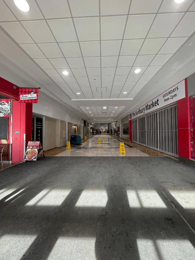 Innenansicht eines Einkaufszentrums mit Deckenbeleuchtung, einem Stuhl und einer Tafel mit der Aufschrift "Sydbury Market - Vorher und Nachher" auf der linken Seite und verstreuten Gegenständen auf der rechten Seite und dem Boden.