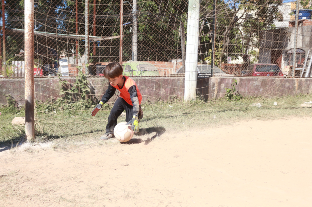 Junge in Fußballschuhen auf einem Schotterfeld mit Gras, Pflanzen, Pfosten, einem Zaun, einer Wand, Bäumen, Fahrzeugen, Gebäuden und einer bewölkten Himmel im Hintergrund.
