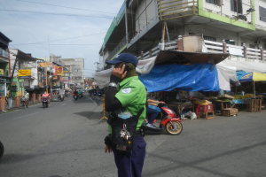 Ein Polizist in grüner und blauer Uniform und blauer Mütze steht an der Seite einer Straße und spricht auf einem Handy, während Fahrzeuge auf der Straße hinter ihm fahren, Stände mit verschiedenen Gegenständen rechts von ihm und ein bewölkter Himmel mit einem Gebäude mit Fenstern im Hintergrund.