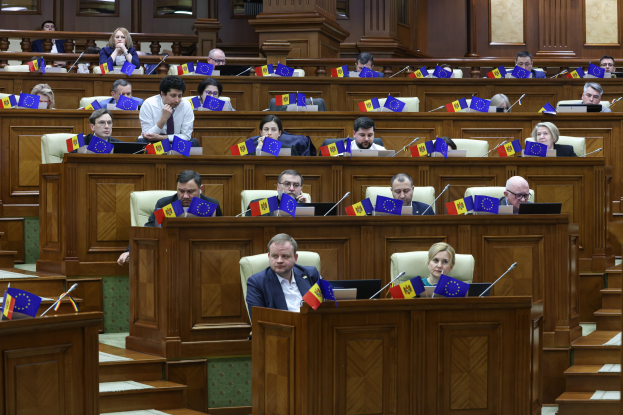 Große Gruppe von Menschen an Tischen in einem Raum mit Flaggen und Mikrofonen, Treppe führt zu den Tischen hoch und Fenster an der Rückwand während einer Sitzung des Europäischen Parlaments.