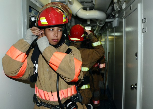 Feuerwehrleute in Uniform während einer Übung in einem Raum stehend, mit einer Tür auf der rechten Seite und sichtbaren Rohren im Hintergrund.