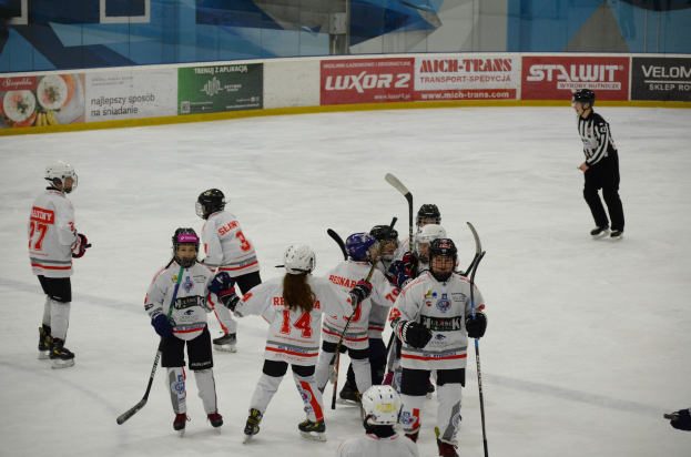 Junge Menschen in Helmen und Hockey-Ausrüstung spielen Eis-hockey auf einem Eisstadion, mit einer Person, die rechts steht und Tafeln mit Text im Hintergrund.