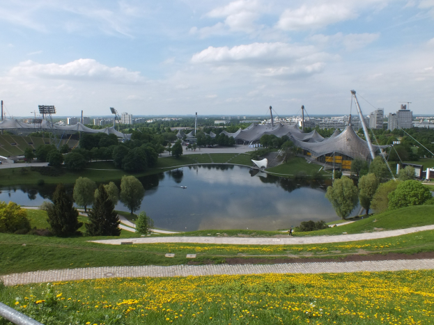 Olympiastadion von einem Hügel aus über einen Teich mit Grünfläche und gelben Blumen, vor einem klaren blauen Himmel mit Gebäuden und Pfählen in der Ferne.