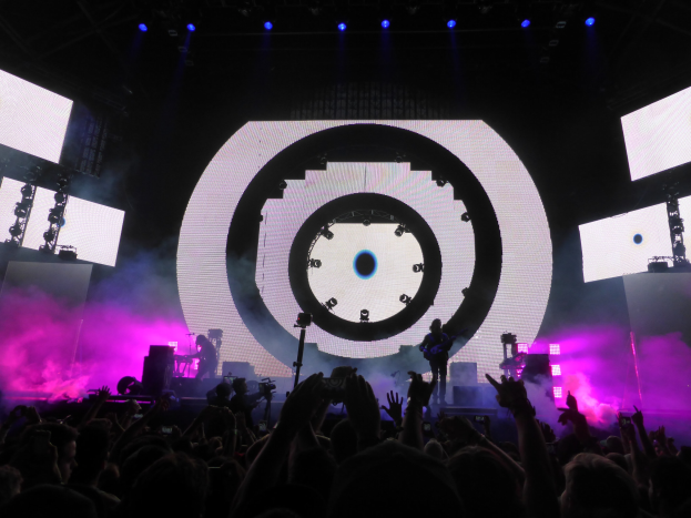 Menge bei einem Konzert mit erhobenen Händen, einige halten Telefone oder spielen Instrumente, großes Display und Lichter im Hintergrund.