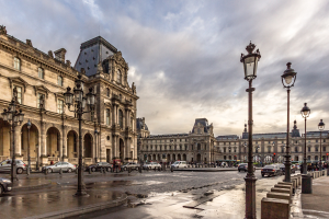 Das Louvre-Museum in Paris, Frankreich, mit seinen ikonischen Gebäuden, Laternenpfählen, Straßenlaternen, Kraftfahrzeugen auf der Straße, Menschen, die auf dem Gehweg gehen, und einem bewölktem Himmel im Hintergrund.