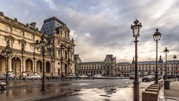 Das Louvre-Museum in Paris, Frankreich, mit seinen ikonischen Gebäuden, Laternenpfählen, Straßenlaternen, Kraftfahrzeugen auf der Straße, Menschen, die auf dem Gehweg gehen, und einem bewölktem Himmel im Hintergrund.