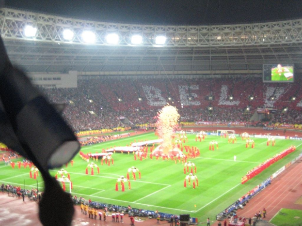 Ein Stadion mit Flutlicht, Eisen Gittern, Zuschauern, stehenden Personen, Werbetafeln, Anzeigetafeln, Überwachungskamera, Fahnen und Dekorationen.