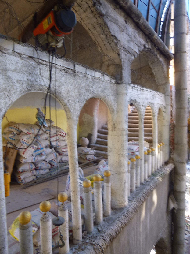 Altes Gebäude mit Bogen, Säulen und Treppen wird renoviert, mit verstreuten Werkzeugen, Materialien und Gegenständen, darunter Taschen und eine Maschine mit Drähten oben drauf.