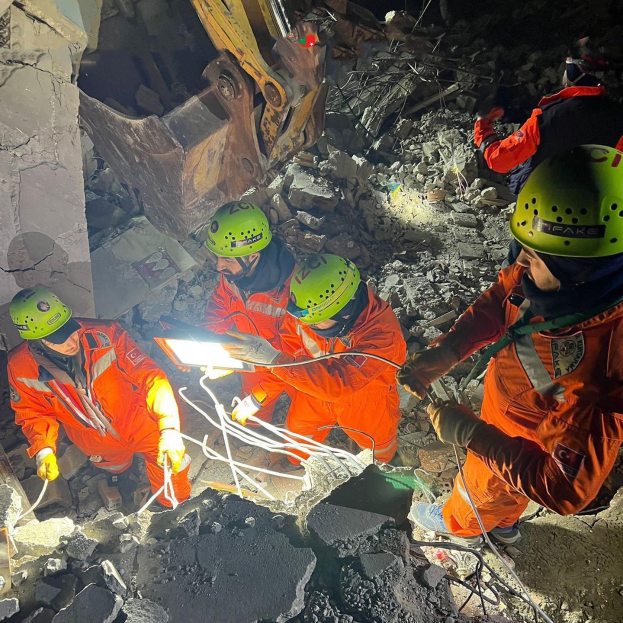 Eine Gruppe von Menschen in orangefarbenen Uniformen und grünen Helmen bei der Arbeit an Trömmern eines eingestürzten Gebäudes, mit einem Kran im Hintergrund.