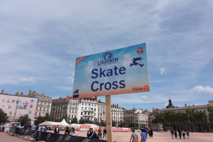 Großes Werbeplakat in einem Stadtplatz umgeben von Passanten, mit Bannern, Zelten, einem Zaun und umliegenden Gebäuden unter einem bewölkten Himmel.