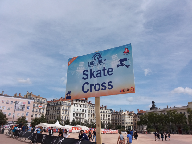 Großes Werbeplakat in einem Stadtplatz umgeben von Passanten, mit Bannern, Zelten, einem Zaun und umliegenden Gebäuden unter einem bewölkten Himmel.