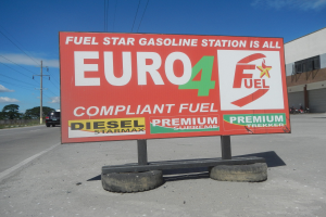 Eine Tankstelle mit einem "Euro4 Fuel"-Schild im Vordergrund, Fahrzeuge auf der Straße, Strommasten mit Drähten, Bäume und ein Gebäude im Hintergrund, unter einem sichtbaren Himmel.