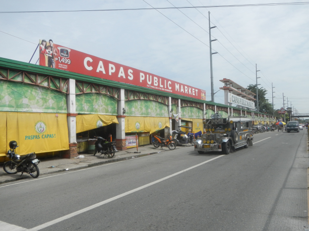 Eine belebte Stadtstraße mit Fahrzeugen, einem Fußweg, Strommasten, Gebäuden, Bäumen und einem bewölkten Himmel, mit einem Gebäude mit der Aufschrift "Capas Public Market" im Vordergrund.