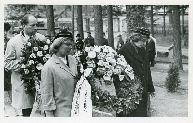 Schwarz-weißes Bild einer Gruppe von Menschen mit Blumensträußen bei einer Gedenkfeier für Holocaust-Opfer, mit Bäumen und Gebäuden im Hintergrund.