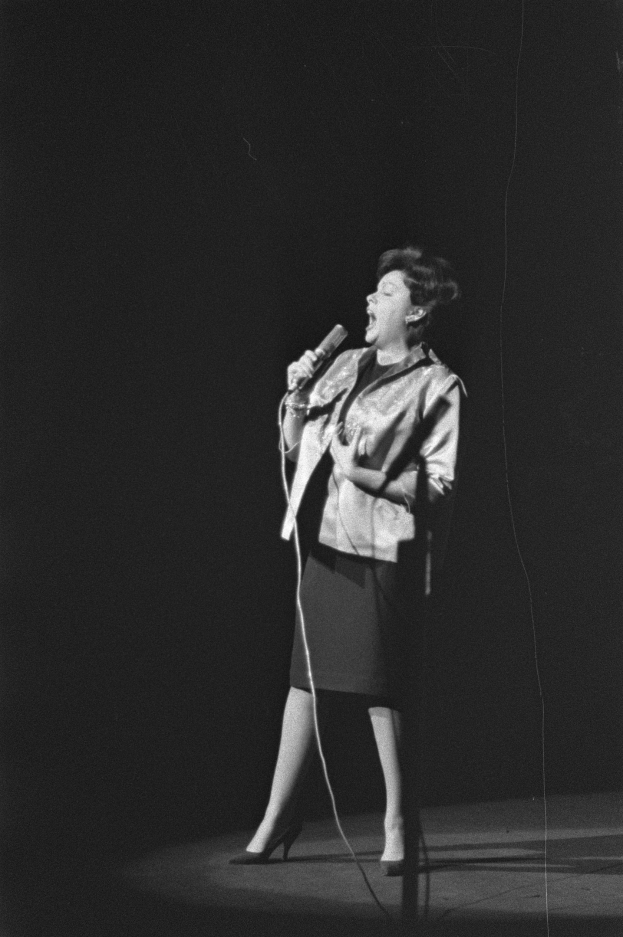 Eine Frau in einem schwarz-weißen Outfit aus den 1950er-Jahren singt fröhlich auf einer von Scheinwerfern beleuchteten Bühne und hält ein Mikrofon in der Hand.