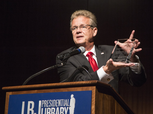 Ein Mann in Anzug und Krawatte, der eine Auszeichnung an einem Podium mit Mikrofon und einer Tafel hält, vor einem dunklen Hintergrund bei einer Bibliothek-Veranstaltung.
