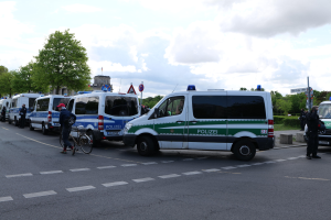 Eine Gruppe von Polizeifahrzeugen steht entlang einer Straße mit einem Radfahrer im Vordergrund und Gebäuden, Bäumen und Beschilderung im Hintergrund.
