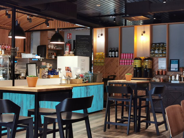 Innenraum eines Restaurants mit Holztischen, Stühlen, einer Topfpflanze auf einem Tisch, einer Tafel, einem Kühlschrank, einem Feuerlöscher, Regalen mit Flaschen, Schränken mit Gegenständen, einem Foto auf der Wand und Deckenleuchten.