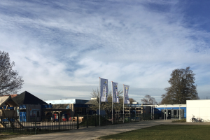 Großes Gebäude mit Metallzaun, Rasenfläche, Weg, Bäumen, Bannern, einem geparkten Fahrrad, nahen Häusern und einem bewölkten Himmel, identifiziert als der neue Spielplatz der Schule.