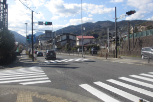 Eine Straßenstadt mit Autos, einer Zebrastreifen, Strommasten, Ampeln, Gebäuden, Bäumen, Hügeln und einem bewölkten Himmel.