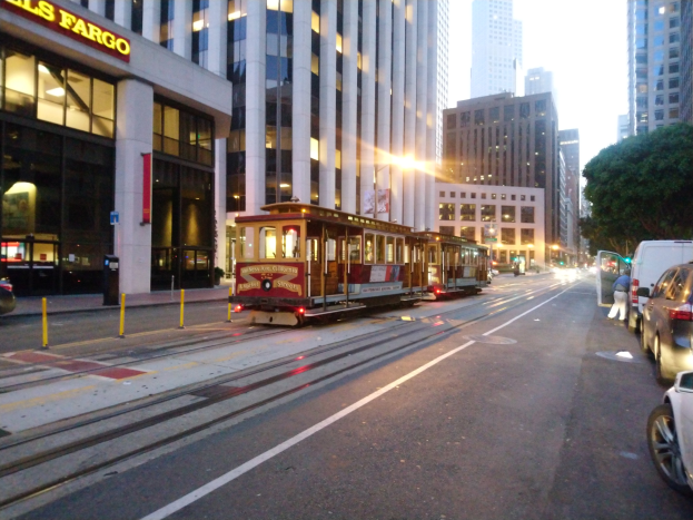 Eine Seilbahn fährt auf einer Straße in San Francisco, mit Fahrzeugen, einem Fußgänger, einem geparkten Van, Gebäuden, Bäumen und Himmel im Hintergrund.