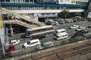 Belebte Stadtstraße mit Fahrzeugen, Gebäuden, Grünanlagen und städtischer Infrastruktur einschließlich Schienen, Geländern und Beschilderung.