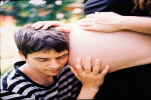 Ein Mann und eine Frau stehen nebeneinander, wobei der Mann zärtlich den Bauch der Frau berührt und auf ihr ungeborenes Kind fokussiert.