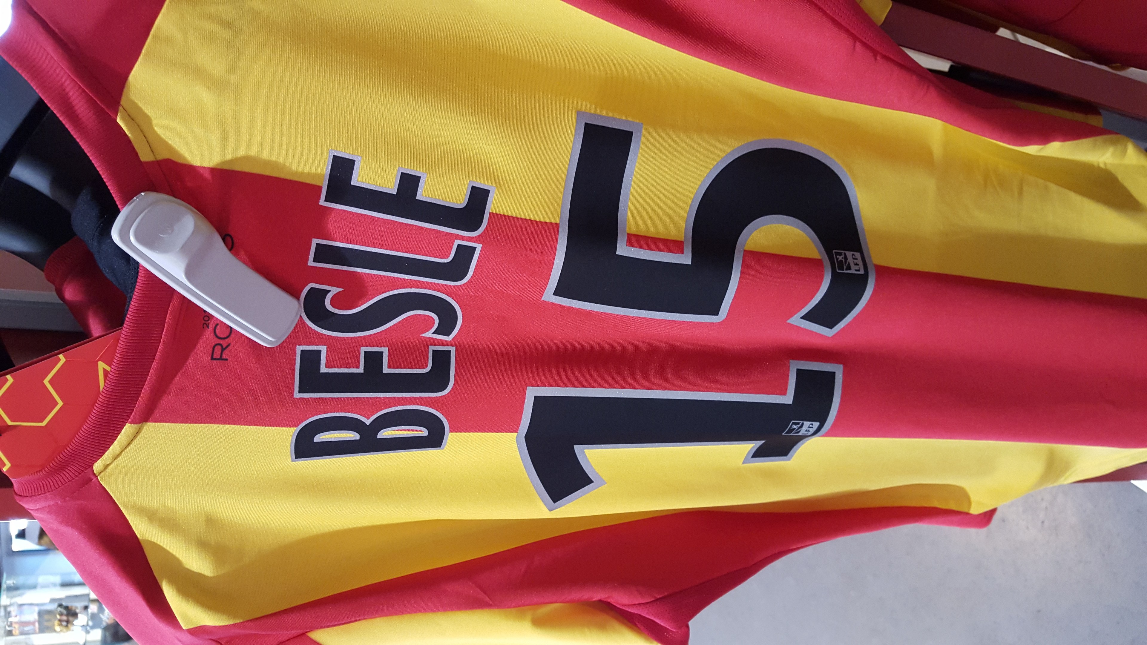 Rotes und gelbes Fußballtrikot mit aufgedrucktem Text und Nummern, das an einer Stange in einem Geschäft hängt, mit anderen Gegenständen und dem Boden im Hintergrund.