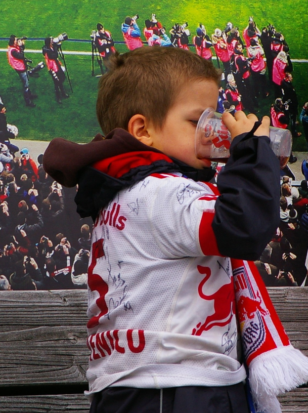 Ein junger Junge trinkt aus einem Glas, während er vor einer Menge mit Kameras steht, mit einer Tafel und Gras im Hintergrund.