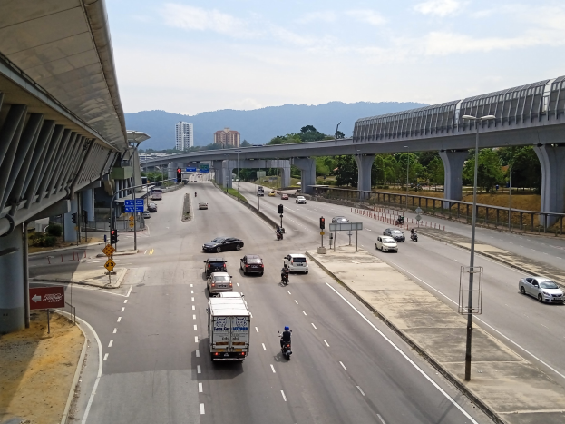 Eine vielbefahrene Autobahn mit mehreren Fahrzeugen in der Nähe einer Brücke, mit Straßenlaternen, Verkehrsampeln, Verkehrszeichen, Bäumen, Gebäuden, Hügeln und einem bewölkten Himmel.