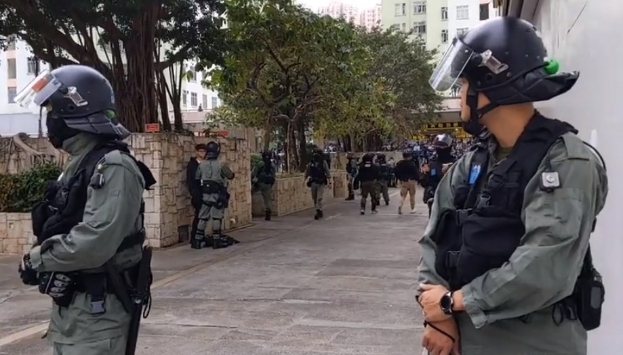 Eine Gruppe von Polizisten in Helmen und kugelsicheren Westen steht auf einer Straße in Hong Kong, mit Bäumen, Gebäuden und Himmel im Hintergrund.