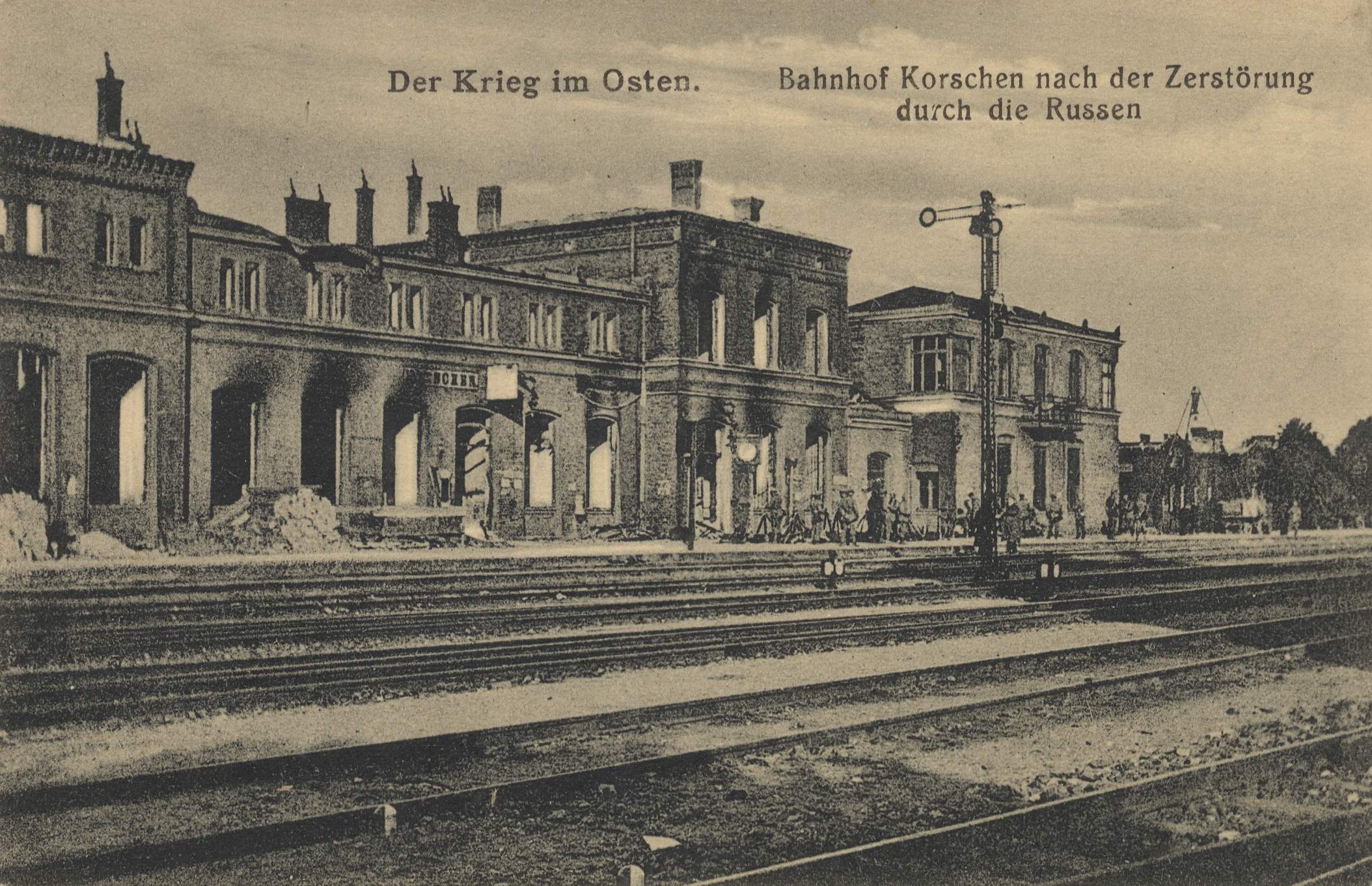 Schwarzes und weißes Foto einer Bahnstation in Korschen Ost, Deutschland, mit Bahnschienen, Gebäuden, Strommasten, Menschen, Bäumen und einem Himmel im Hintergrund.
