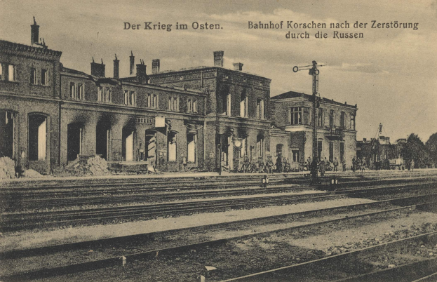 Schwarzes und weißes Foto einer Bahnstation in Korschen Ost, Deutschland, mit Bahnschienen, Gebäuden, Strommasten, Menschen, Bäumen und einem Himmel im Hintergrund.