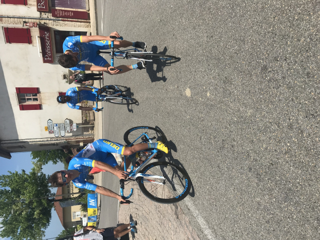 Eine Gruppe von Radfahrern fährt eine Straße entlang an einem Gebäude mit Fenstern und Texttafeln vorbei, mit Bäumen und einem blauen Himmel im Hintergrund und Zuschauern auf der Straße.