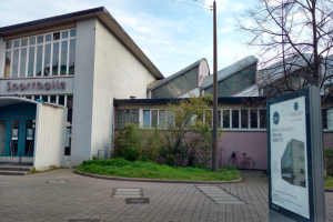 Ein großes Gebäude mit einem Schild davor, umgeben von Bäumen, Pflanzen, Gras, Fahrrädern und einem Pfahl, mit einem bewölkten Himmel im Hintergrund, identifiziert als Sportballe, geplant zur Eröffnung im Jahr 2020.