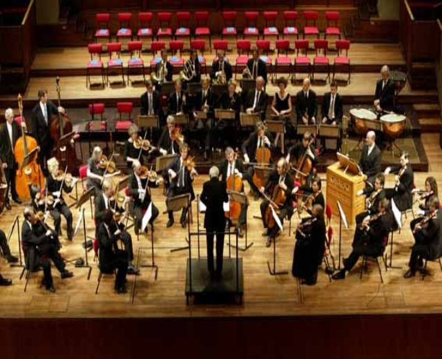 Großes Orchester auf der Bühne mit dirigierendem Musiker, Musikständer und Hintergrundtreppe.