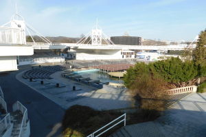 Ein Blick auf das Olympische Stadion von einem hohen Punkt aus, mit verschiedenen Objekten und Grünflächen im Vordergrund und einem bewölkten Himmel im Hintergrund.