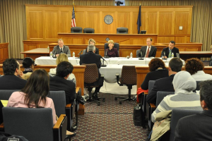 Ein Tagungsraum mit einer Gruppe von Menschen, die auf Stühlen sitzen, zwei ausländischen Flaggen, einem orangen Schreibtisch mit Wasserflaschen und einem Mikrofon.