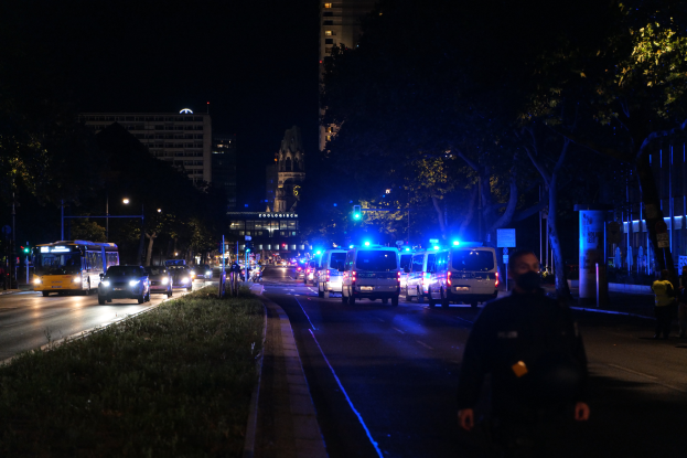 Eine nächtliche Straßen Szenerie mit Polizeiwagen, Bussen, Fußgängern auf dem Gehweg, Gras, Bäumen, Gebäuden und beleuchteten Laternen.