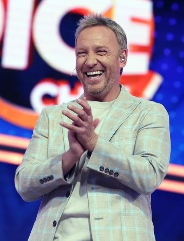 Ein Mann im Anzug klatscht auf der Bühne mit 'The Voice'-Logo im Hintergrund.