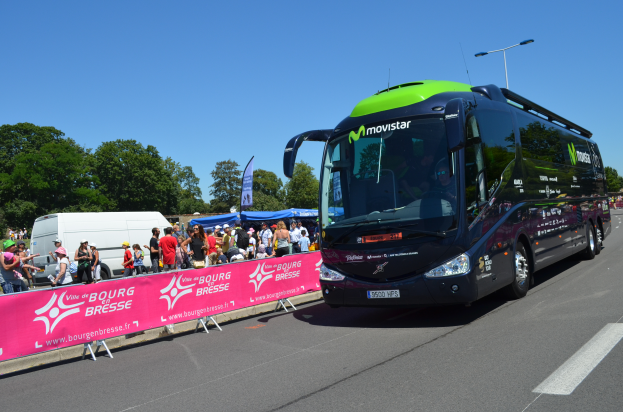 Ein schwarzer und grüner Bus fährt auf einer Straße neben einer Menge Menschen, einige tragen Möppen, mit einem Banner auf der linken Seite und Bäumen unter einem klaren blauen Himmel im Hintergrund.