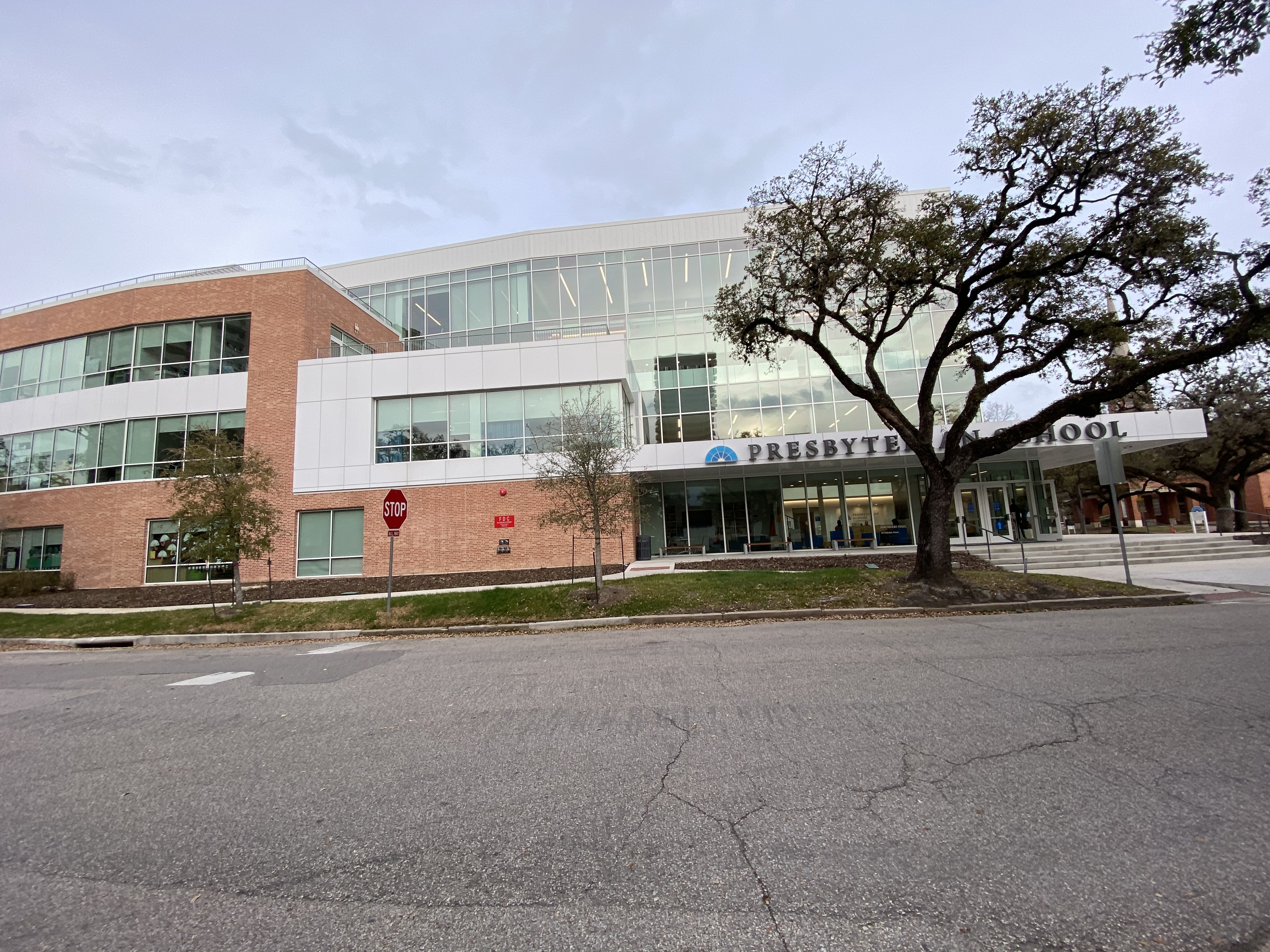 Presbyte Mittelschule, ein großes Gebäude mit vielen Fenstern, umgeben von Bäumen, Gras, Schildern, Pfosten und einer Straße, mit einem bewölkten Himmel im Hintergrund.