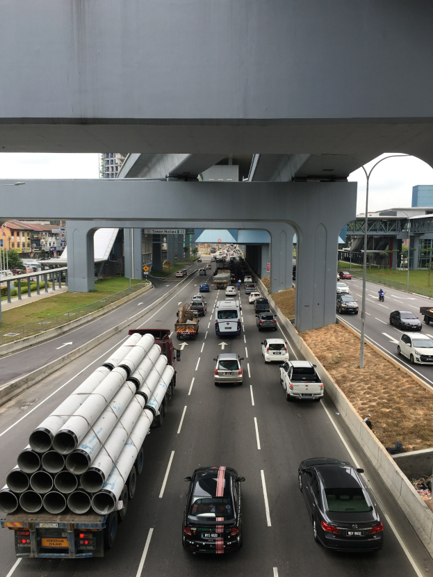 Eine befahrene Autobahn mit mehreren Fahrzeugen, eine Brücke darüber, Straßenlaternen, Gras, Gebäude und Bäume unter einem klaren Himmel.