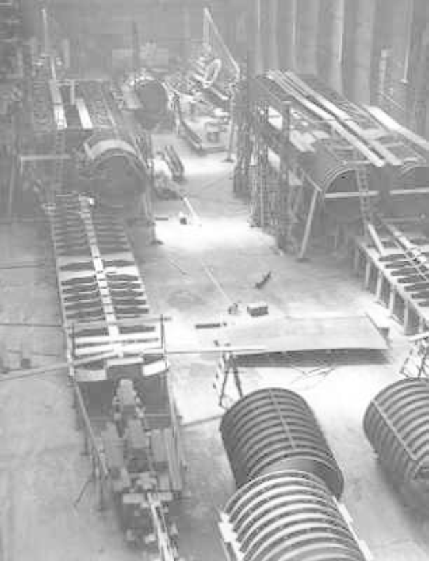 Schwarzes und weißes Foto eines industriellen Fabrikinneren mit zahlreichen Rohren, Maschinen und Geräten auf dem Boden, vor strukturellen Säulen und einer Wand.